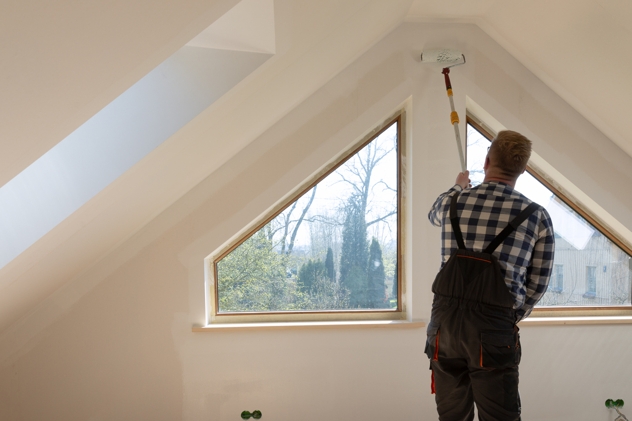Home improvement concept, handyman painting a wall with a white paint near roof window in attic